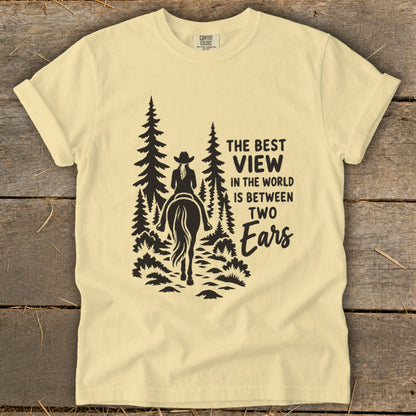 A light yellow t-shirt with a graphic of a horseback rider in pine trees and the text THE BEST VIEW IN THE WORLD IS BETWEEN TWO EARS.