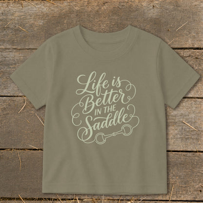 Tan t-shirt with "Life is Better IN THE Saddle" text and horse bit graphic.