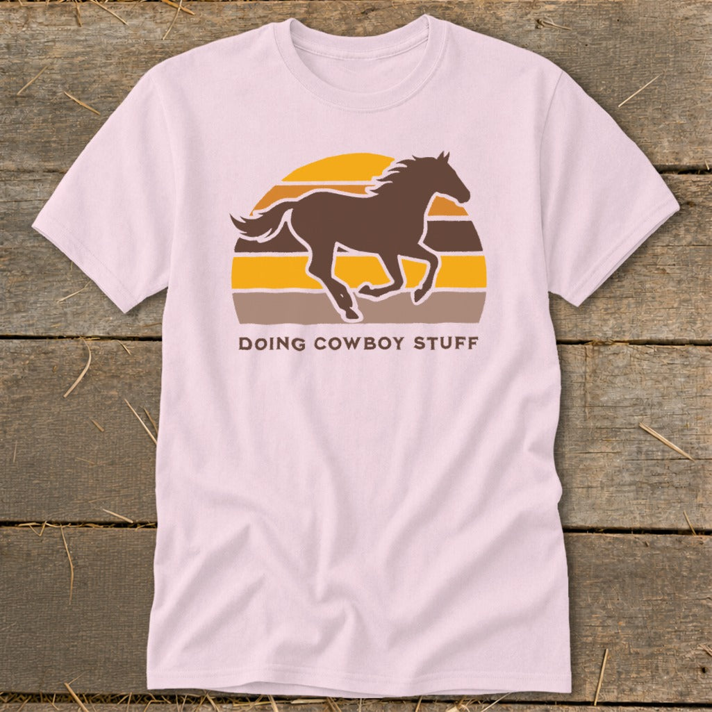 Light pink t-shirt with a brown horse running in front of a sunset and the text "DOING COWBOY STUFF".