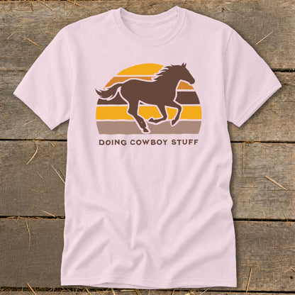 Light pink t-shirt with a brown horse running in front of a sunset and the text "DOING COWBOY STUFF".