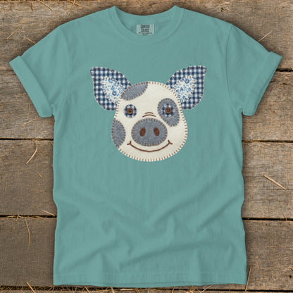 Teal t-shirt with a fabric appliqué of a pig's face with blue checkered ears.