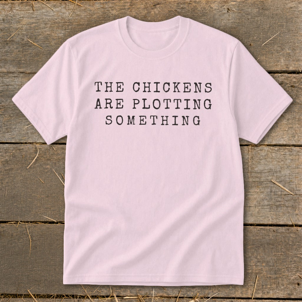 Light pink t-shirt with black text that reads "THE CHICKENS ARE PLOTTING SOMETHING".