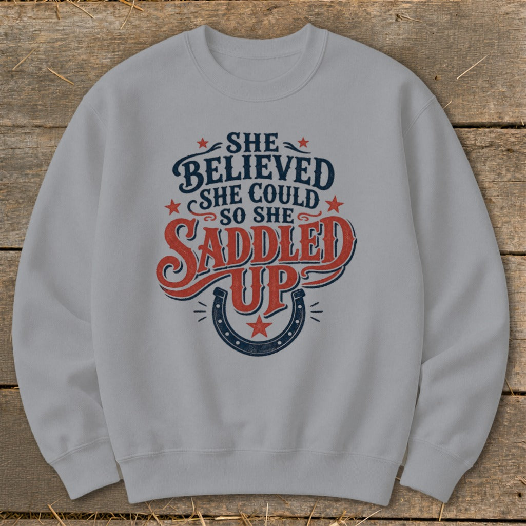 Grey sweatshirt with "She Believed She Could So She Saddled Up" text and horseshoe graphic.