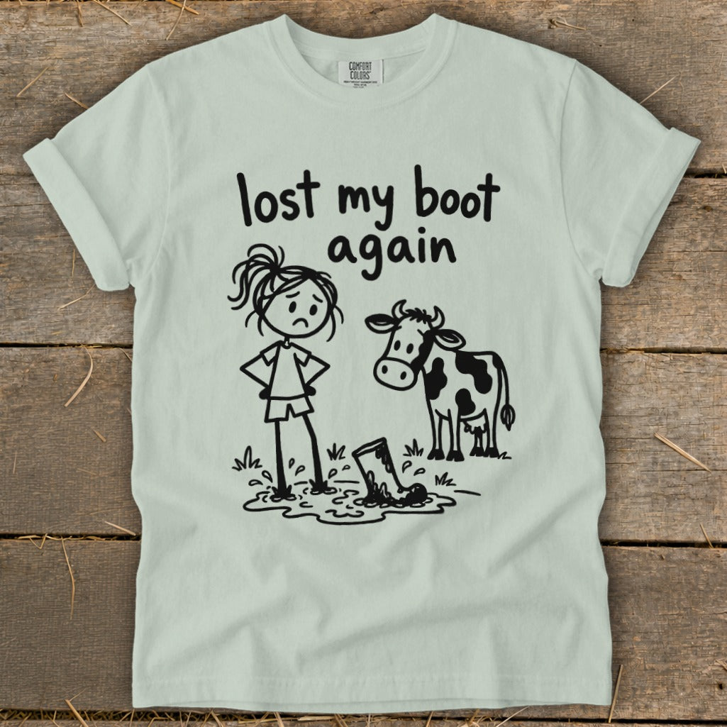 Light green t-shirt featuring a cartoon of a girl in mud next to a cow, with the text "lost my boot again".