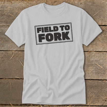 A light gray t-shirt featuring the black text "FIELD TO FORK" inside a rectangular border, laid flat on a wooden surface.