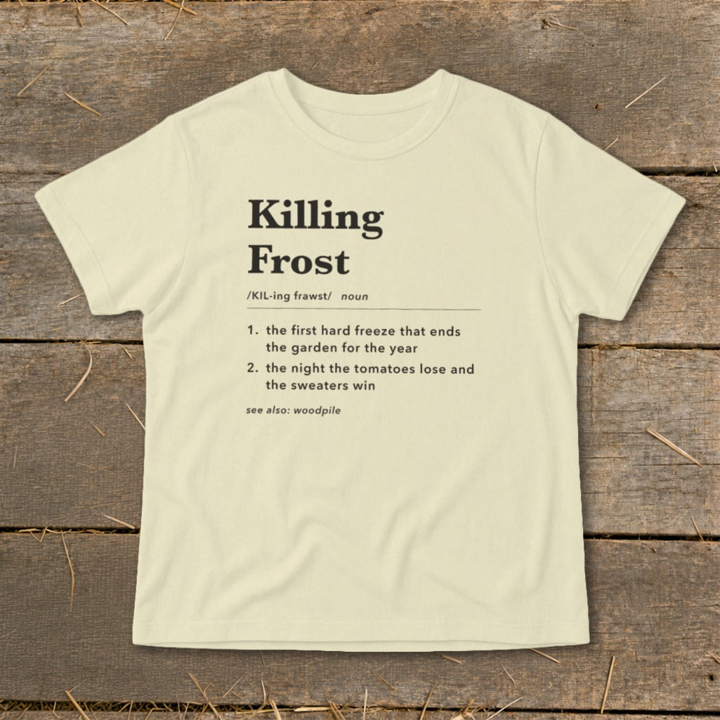 A cream-colored t-shirt with black text featuring a humorous definition of Killing Frost laid flat on wooden planks.