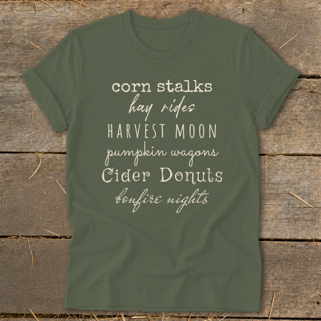 Green t-shirt with "corn stalks hay rides harvest moon pumpkin wagons cider donuts bonfire nights" text.