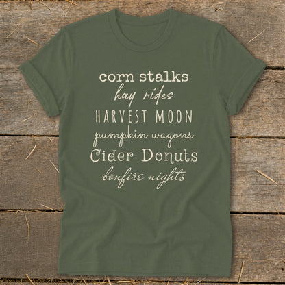 Green t-shirt with "corn stalks hay rides harvest moon pumpkin wagons cider donuts bonfire nights" text.