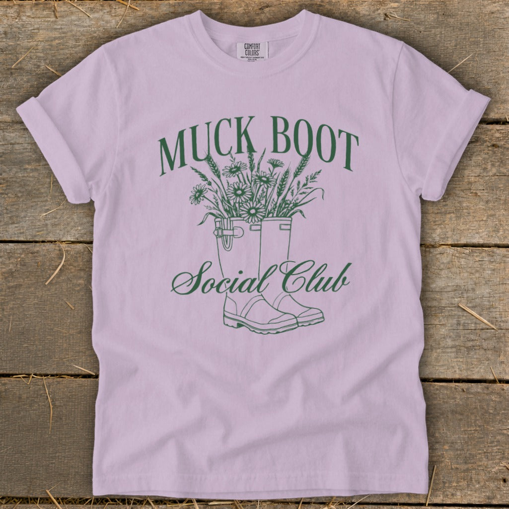 Lavender t-shirt with green graphic of muck boots filled with flowers and text.
