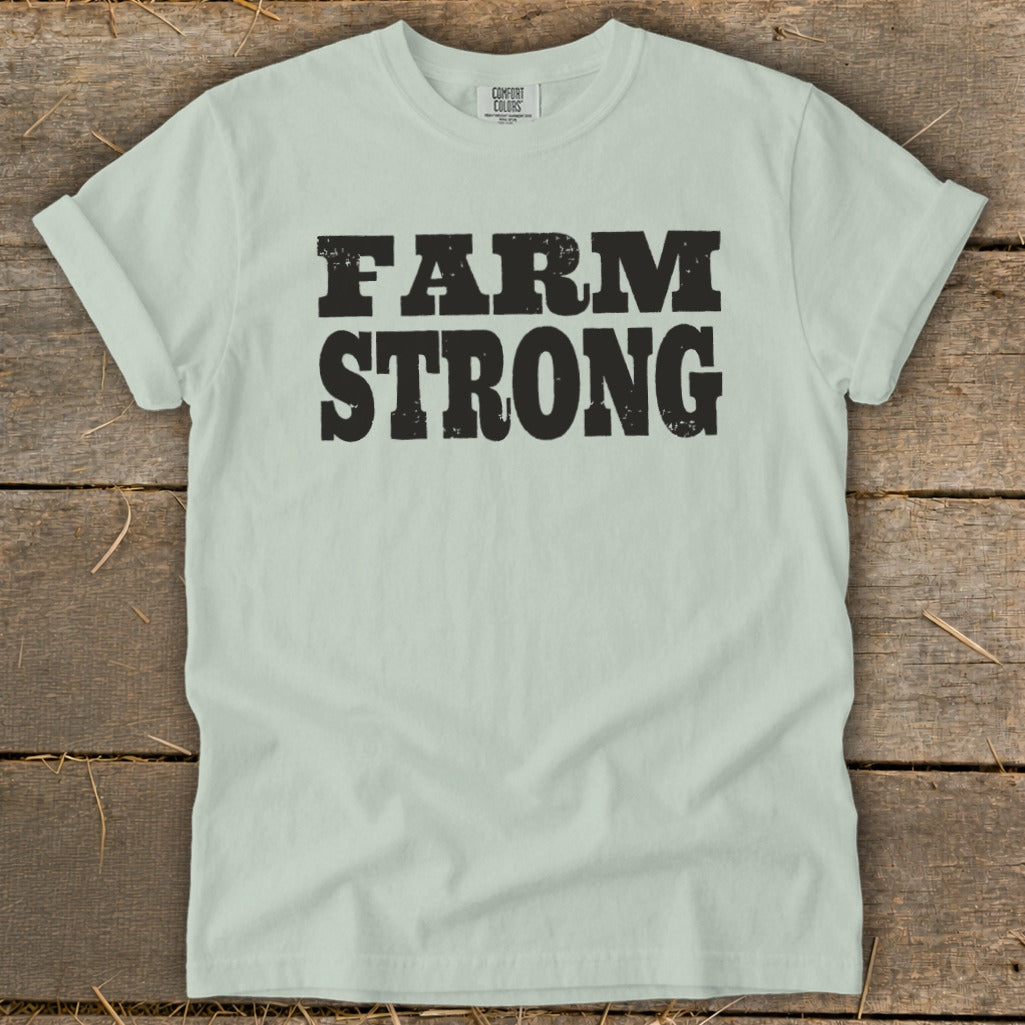 A light green crewneck t-shirt with rolled sleeves featuring the text FARM STRONG in black block letters, laid flat on a rustic wooden background.