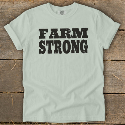 A light green crewneck t-shirt with rolled sleeves featuring the text FARM STRONG in black block letters, laid flat on a rustic wooden background.