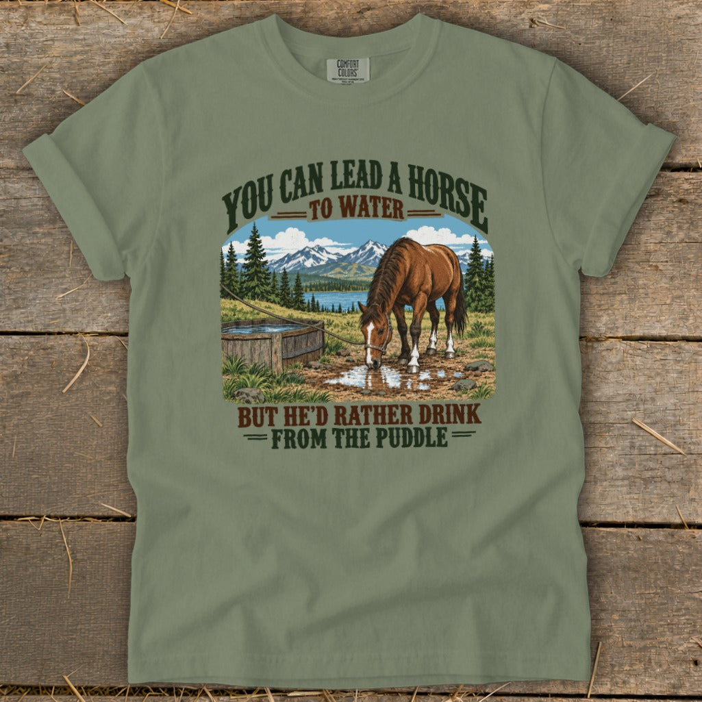 A sage green t-shirt with a graphic of a horse drinking from a puddle with mountains and text.