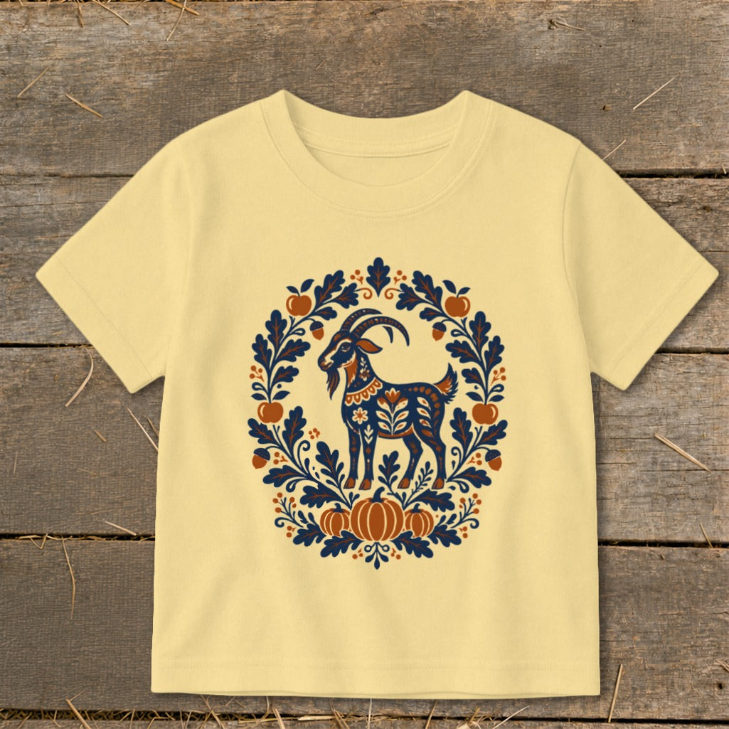 A light yellow t-shirt features a folk-art graphic of a goat surrounded by autumn leaves and pumpkins.