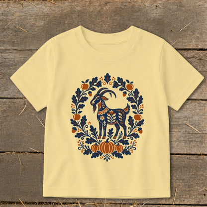A light yellow t-shirt features a folk-art graphic of a goat surrounded by autumn leaves and pumpkins.