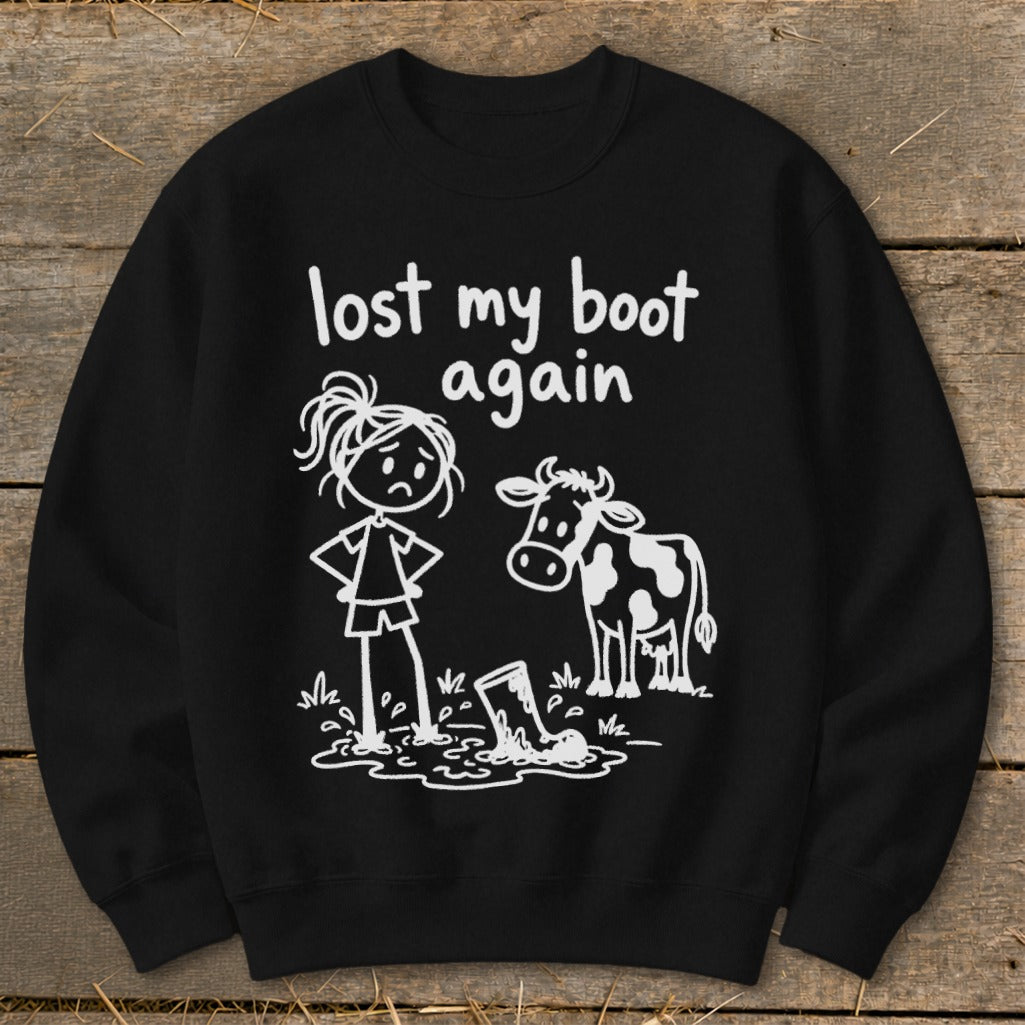Black crewneck sweatshirt printed with white text reading lost my boot again, featuring a stick figure and a cow looking at a muddy boot.