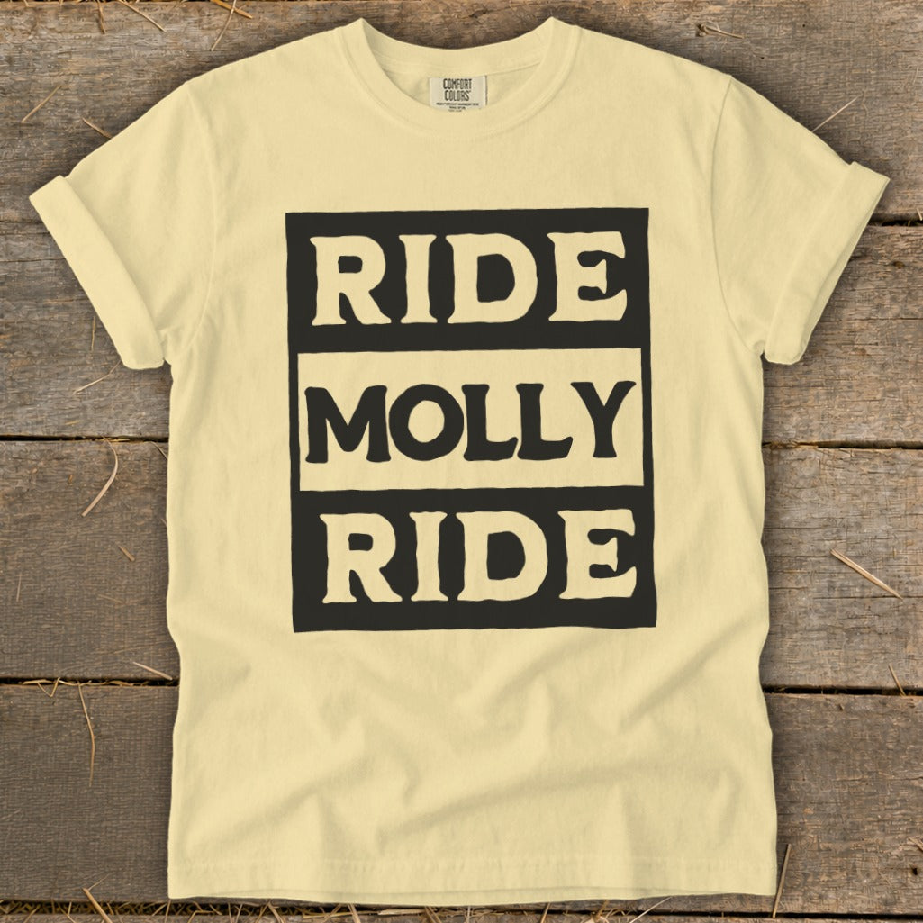 A pale yellow crewneck t-shirt with black text reading Ride Molly Ride on a wooden background.