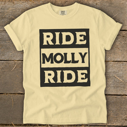 A pale yellow crewneck t-shirt with black text reading Ride Molly Ride on a wooden background.