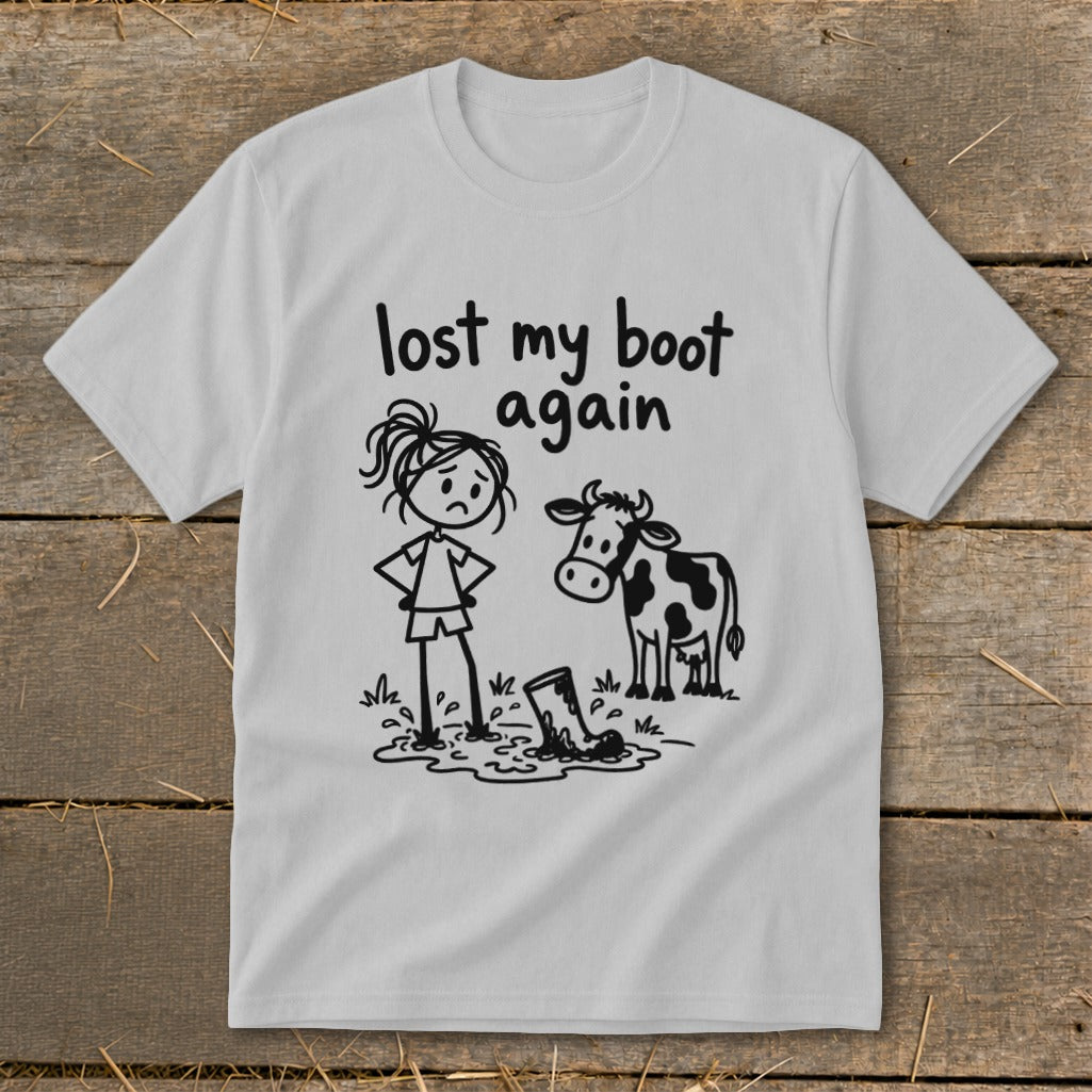 Light gray t-shirt with a black line-drawing graphic of a sad girl standing next to a cow and a boot stuck in mud, accompanied by the text "lost my boot again".