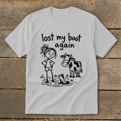 Light gray t-shirt with a black line-drawing graphic of a sad girl standing next to a cow and a boot stuck in mud, accompanied by the text "lost my boot again".