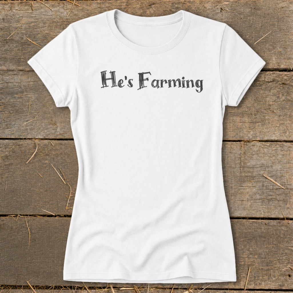 White t-shirt with black text reading "He's Farming" on a wooden background.
