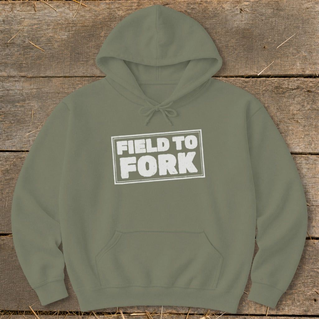 An olive green hooded sweatshirt lies flat on a wooden surface, featuring the white text "FIELD TO FORK" inside a distressed rectangular box on the chest.