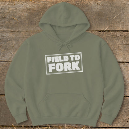 An olive green hooded sweatshirt lies flat on a wooden surface, featuring the white text "FIELD TO FORK" inside a distressed rectangular box on the chest.