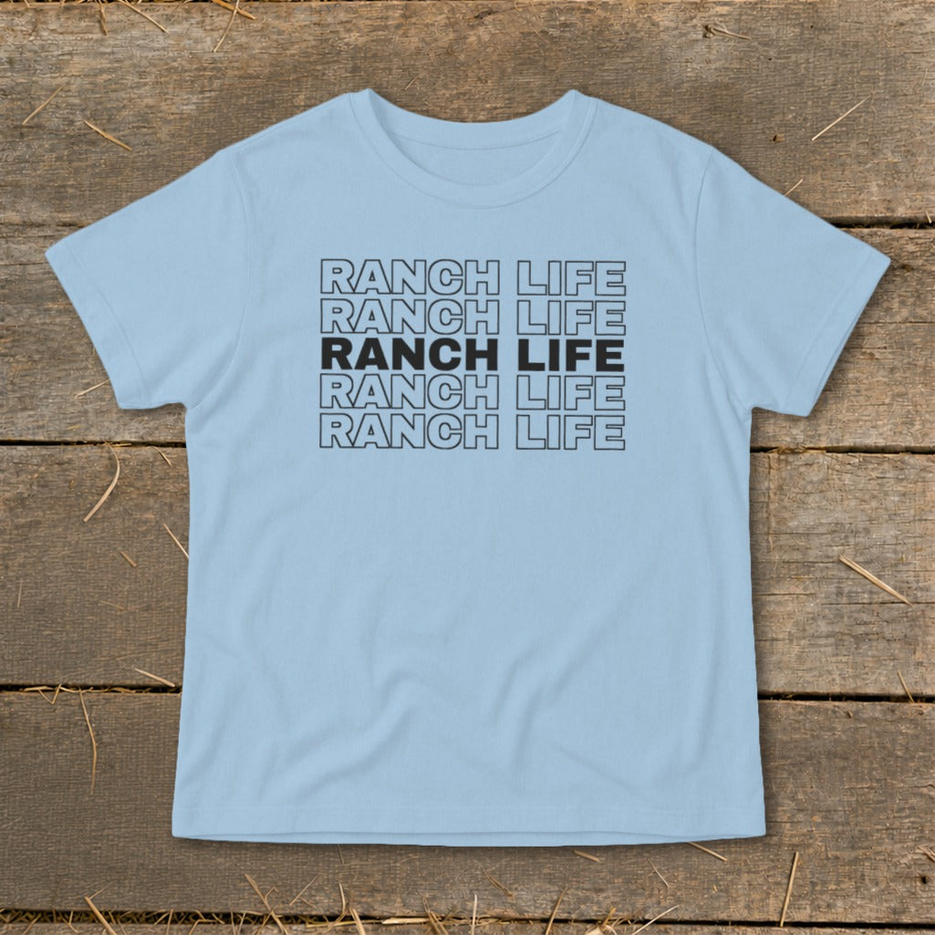 Light blue short-sleeved t-shirt with repeated stacked black text reading "RANCH LIFE" on a rustic wooden background.