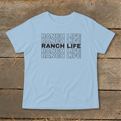 Light blue short-sleeved t-shirt with repeated stacked black text reading "RANCH LIFE" on a rustic wooden background.