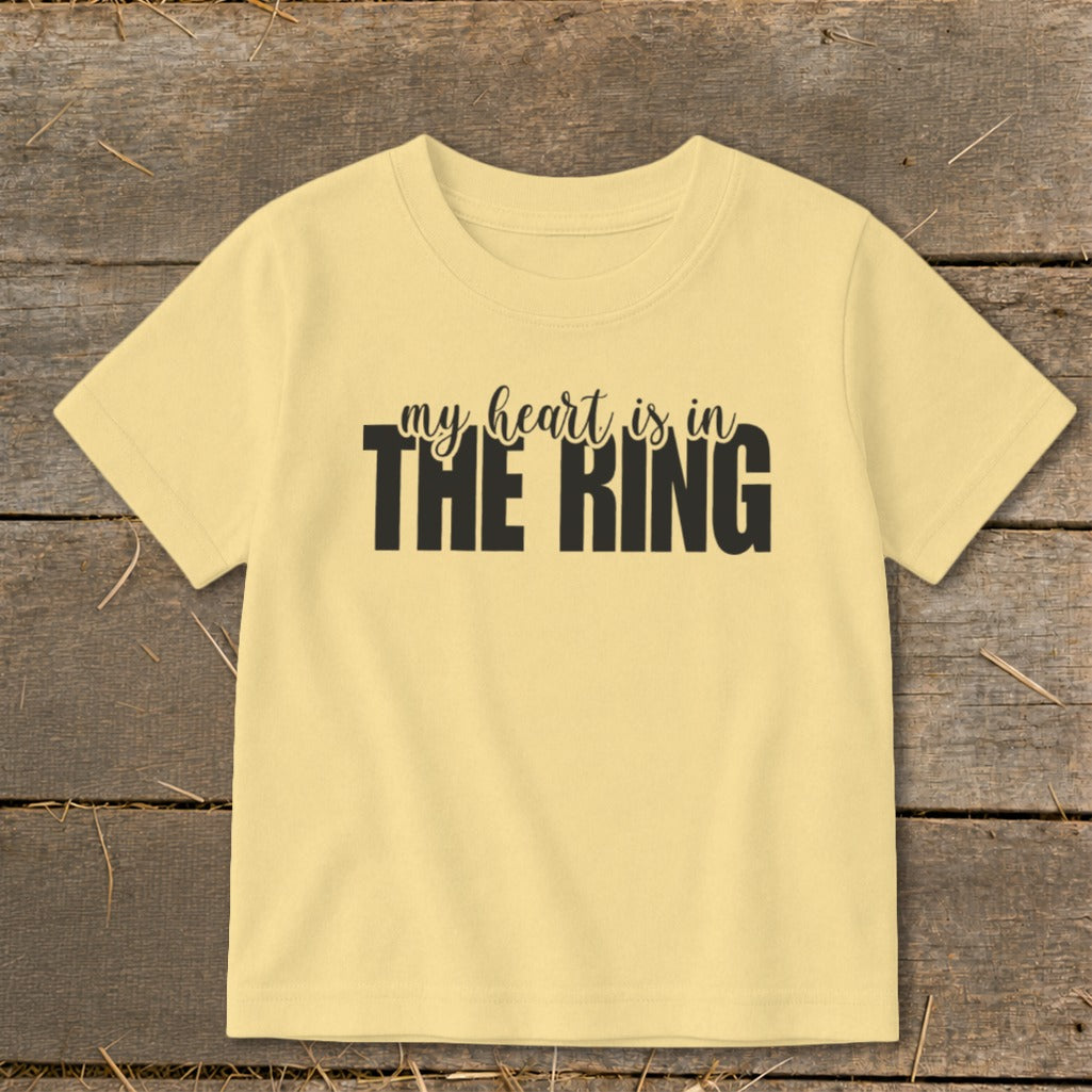 A yellow short-sleeve t-shirt with black text that reads "my heart is in THE RING" centered on the chest, lying flat on a wooden surface.