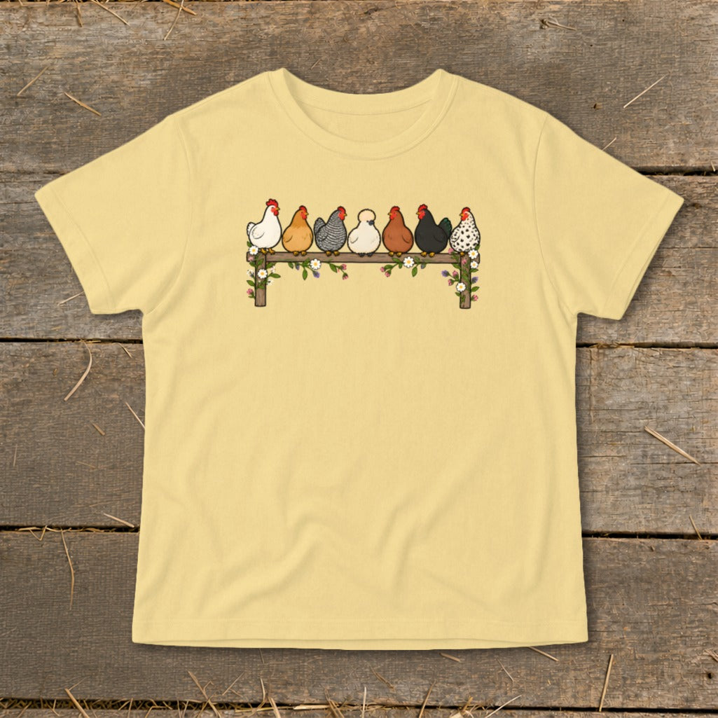 A pale yellow t-shirt features a graphic of seven varied chickens perched on a flower-adorned wooden fence.