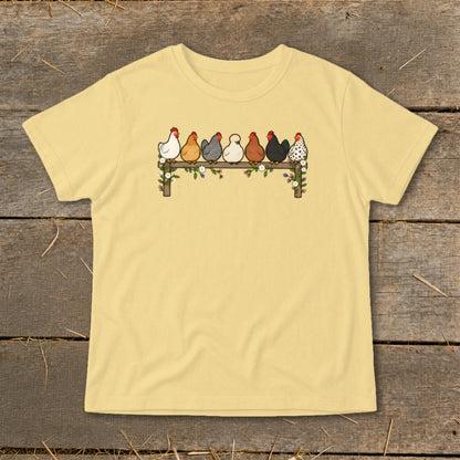 A pale yellow t-shirt features a graphic of seven varied chickens perched on a flower-adorned wooden fence.
