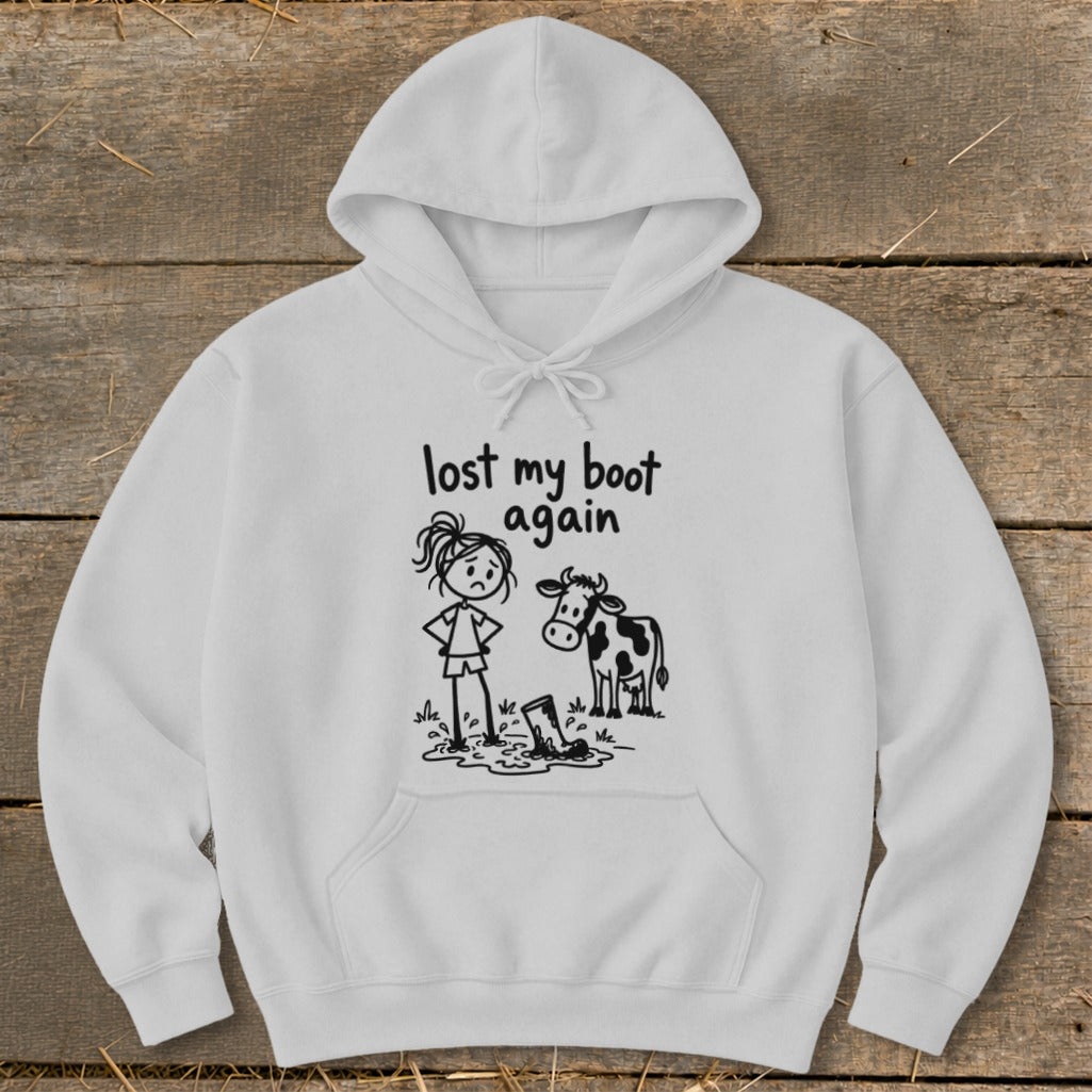 A light gray pullover hoodie featuring a black graphic of a sad stick figure standing next to a boot stuck in mud, accompanied by a curious cow and the text "lost my boot again."