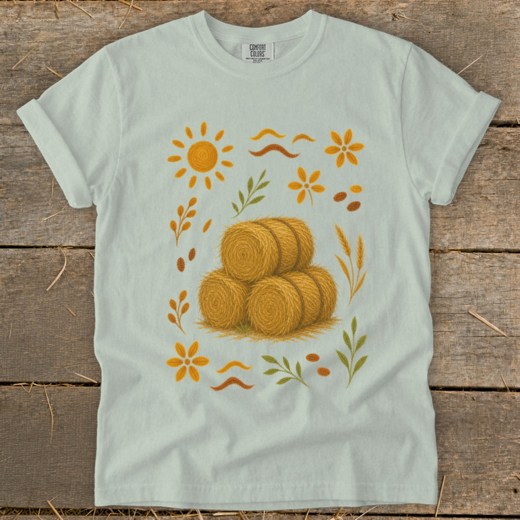 Light blue t-shirt with a graphic featuring hay bales, sun, and floral elements.