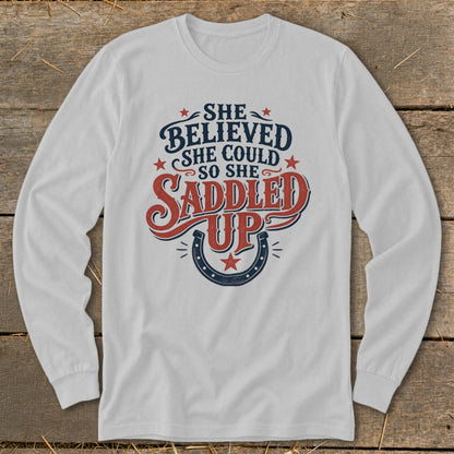 White long-sleeve shirt with "She believed she could so she saddled up" graphic.