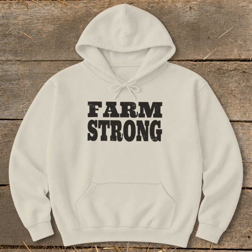 Cream-colored hooded sweatshirt with a front pocket and black distressed text reading FARM STRONG on a wooden background.