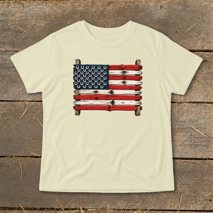 Light-colored t-shirt with a graphic of an American flag made of weathered wood and horseshoes, displayed on wooden planks.
