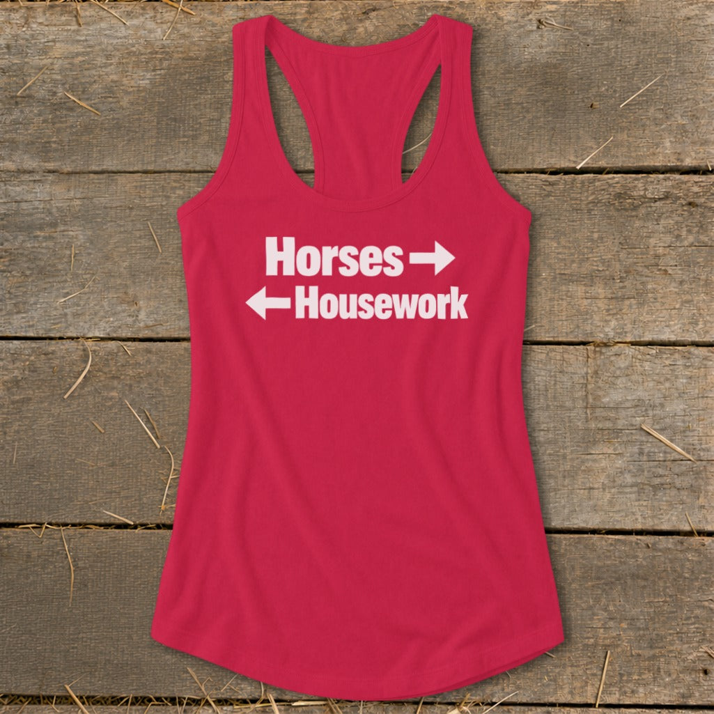 A red women's racerback tank top with white text that reads Horses with a right arrow and Housework with a left arrow, displayed on a rustic wooden background.