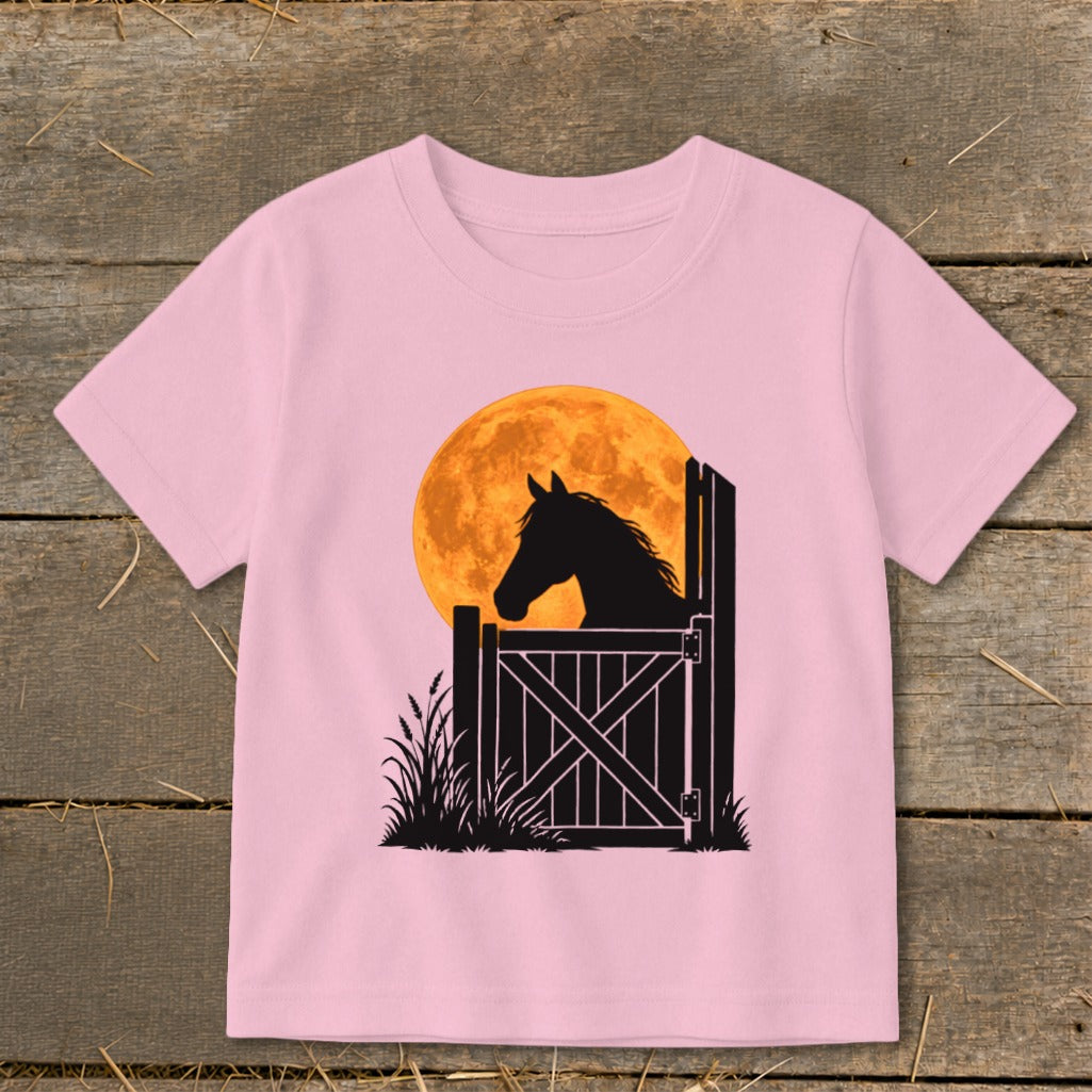 Light pink short-sleeve children's t-shirt featuring a black silhouette graphic of a horse behind a gate against an orange full moon.