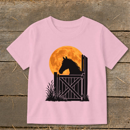 Light pink short-sleeve children's t-shirt featuring a black silhouette graphic of a horse behind a gate against an orange full moon.