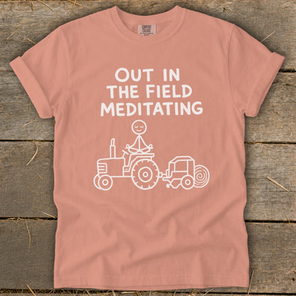 Peach t-shirt with "Out in the Field Meditating" graphic of stick figure on tractor.