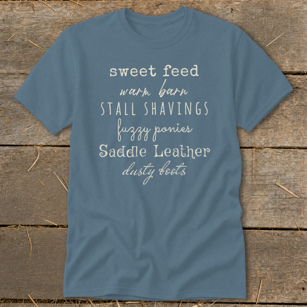 Blue t-shirt with text: sweet feed warm barn stall shavings fuzzy ponies saddle leather dusty boots.