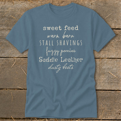 Blue t-shirt with text: sweet feed warm barn stall shavings fuzzy ponies saddle leather dusty boots.