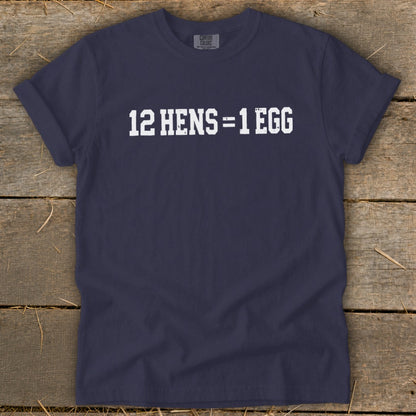 Dark blue t-shirt featuring the white distressed text 12 HENS = 1 EGG, laid flat on a rustic wooden surface.
