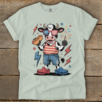 Cartoon cow wearing sunglasses and holding a hot dog, celebrating Fourth of July.