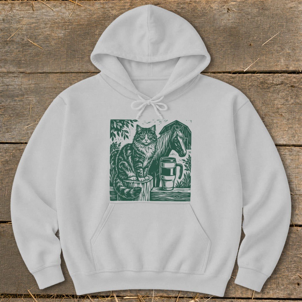 White hoodie with a green linocut design of a cat, horse, and mug on a wooden background.