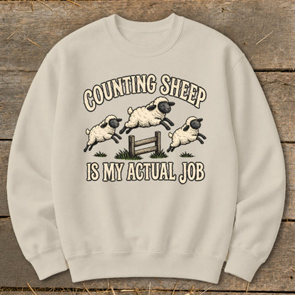 Cream sweatshirt with "Counting Sheep Is My Actual Job" text and jumping sheep graphic.