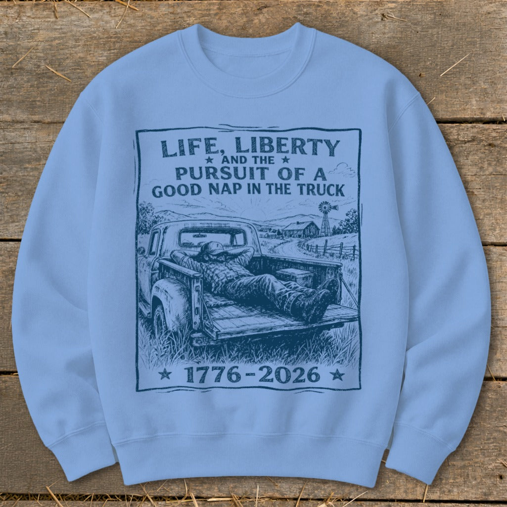 Light blue sweatshirt with a sketch of a man napping in a truck bed. Text reads "Life, Liberty And The Pursuit Of A Good Nap In The Truck 1776-2026".