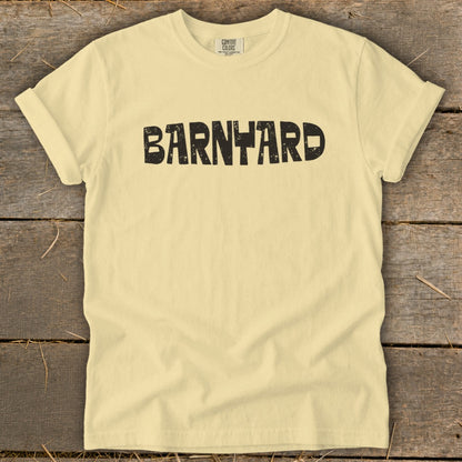 Cream-colored t-shirt with black distressed text reading "BARNYARD" centered on the chest, laid flat on a wooden background.