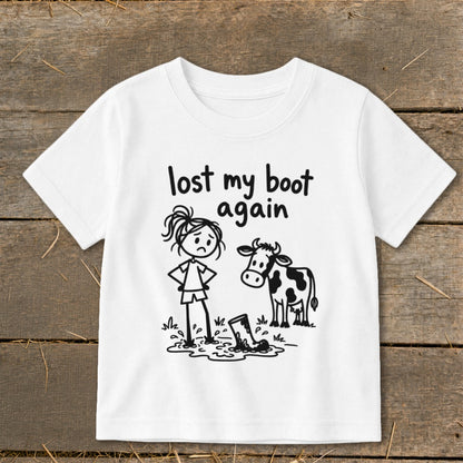 A white short-sleeve t-shirt features a graphic of a sad stick figure standing next to a cow and a muddy boot, with text reading "lost my boot again".