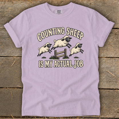 Lilac t-shirt with graphic of three sheep jumping over a fence with text "COUNTING SHEEP IS MY ACTUAL JOB".
